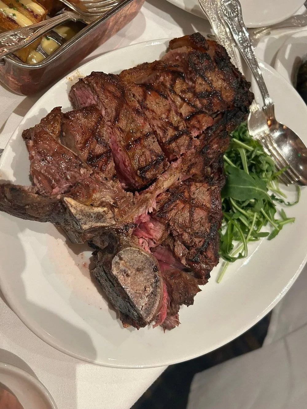A large bone-in T-bone steak, charred from the grill bars, filling a white plate beside a pile of watercress, with silver cutlery and a dish of chips on a white tablecloth
