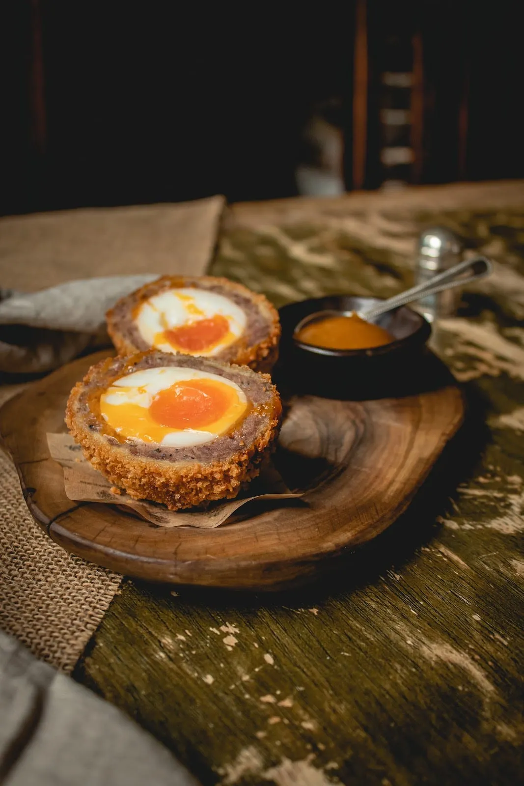 A Scotch egg cut in half, showing a soft egg yolk inside sausage meat and a breadcrumb coating, served with a small pot of sauce.