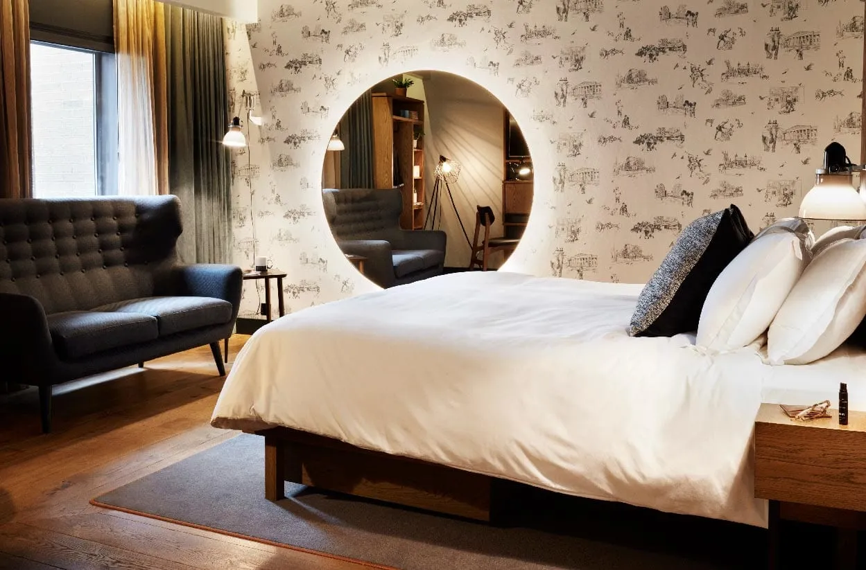 A guest room at the Hoxton Holborn, with illustrated toile wallpaper, a round backlit mirror and a dark blue sofa