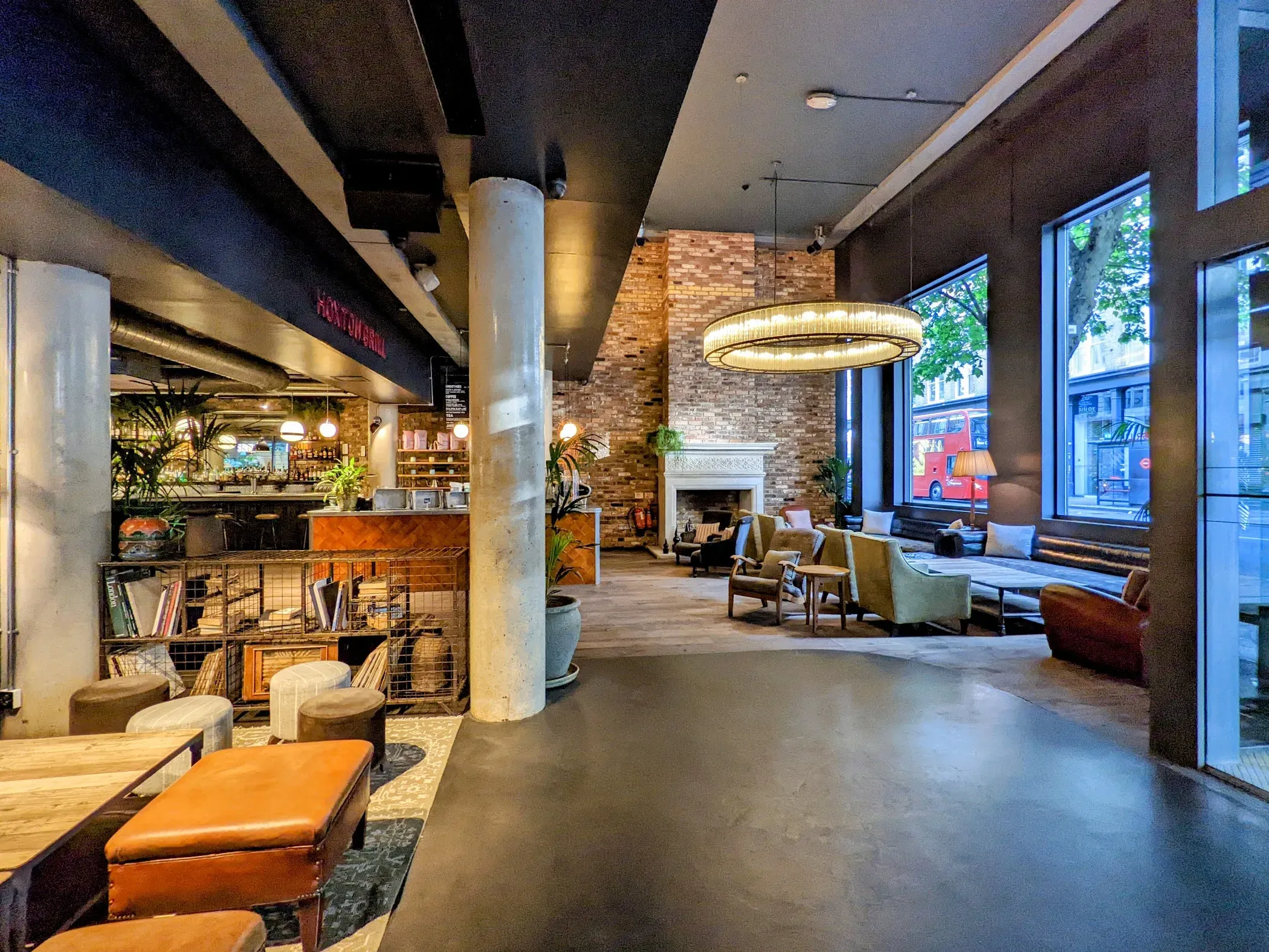 The open lobby at The Hoxton, Shoreditch: a brass ring chandelier over armchairs, an exposed brick fireplace, a herringbone bar under a Hoxton Grill sign, and a red bus passing the window