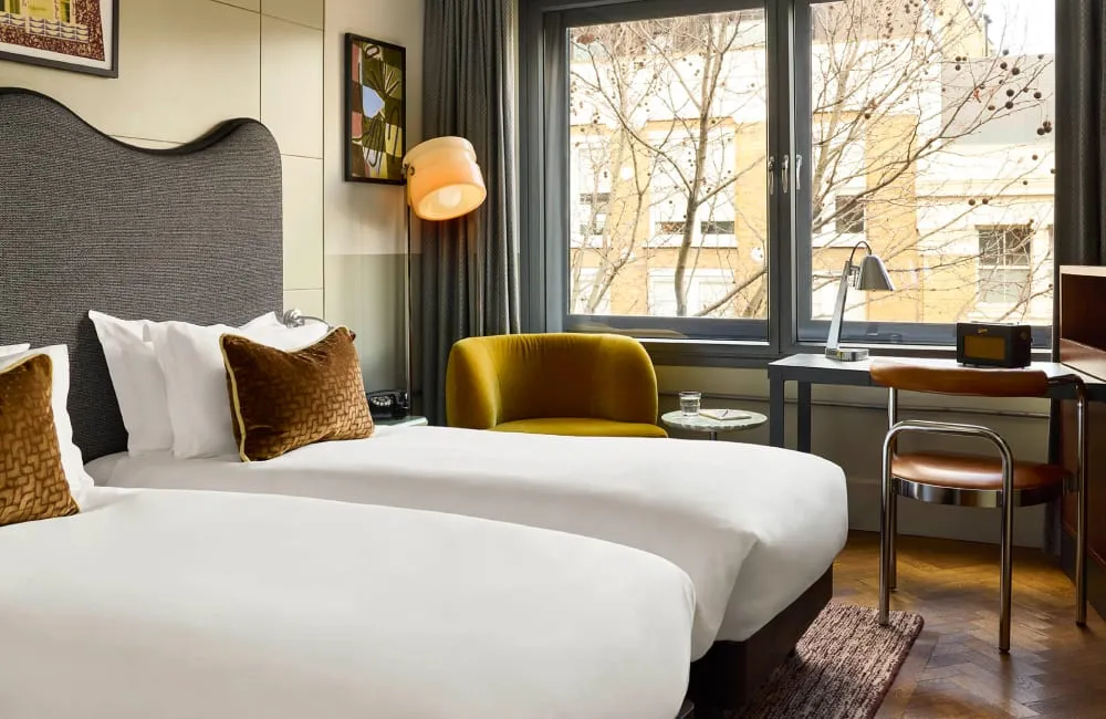 A twin room at The Hoxton, Shoreditch, with a mustard velvet armchair, a curved grey headboard and a writing desk with a retro-style radio
