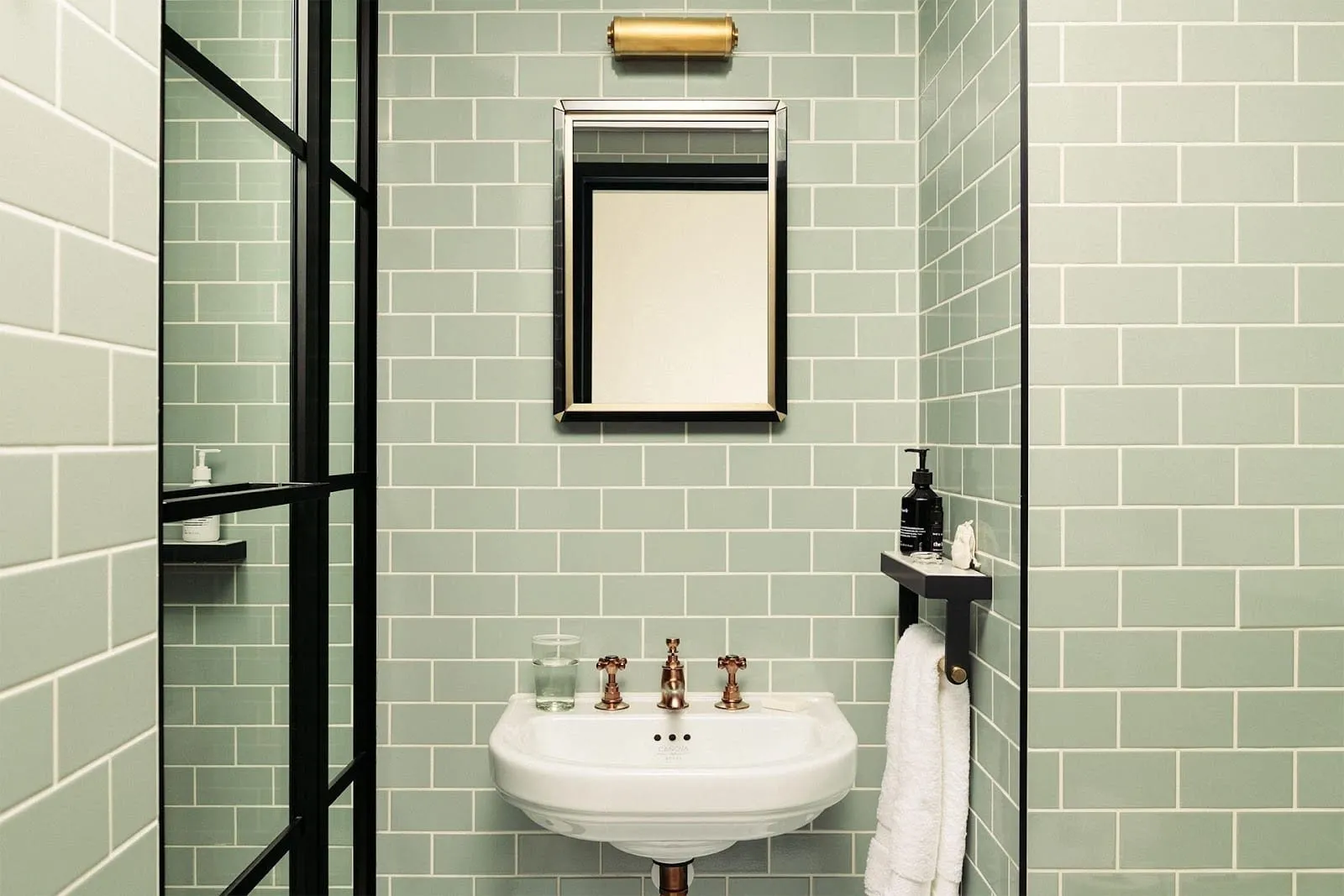 A bathroom at The Hoxton, Southwark with sage-green metro tiles, a pedestal basin with copper cross-head taps and a black-framed glass shower screen