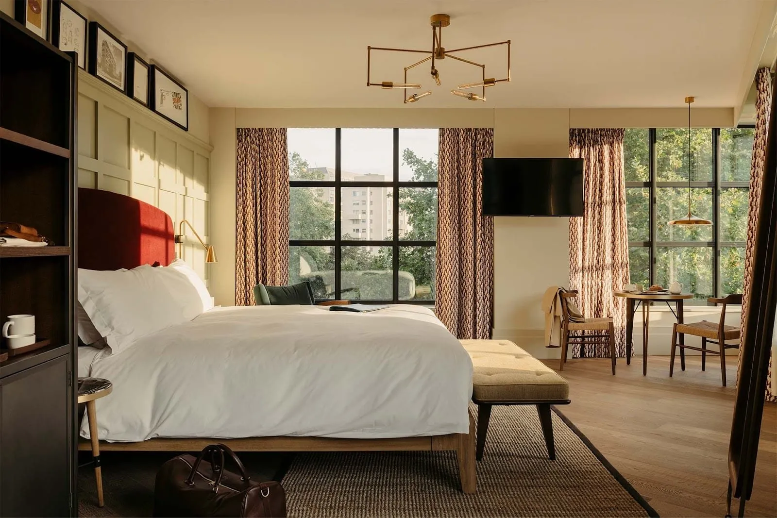 A large room at The Hoxton, Southwark with a red velvet headboard against cream panelling, black-framed factory windows onto trees, a bench at the foot of the bed and a table for two