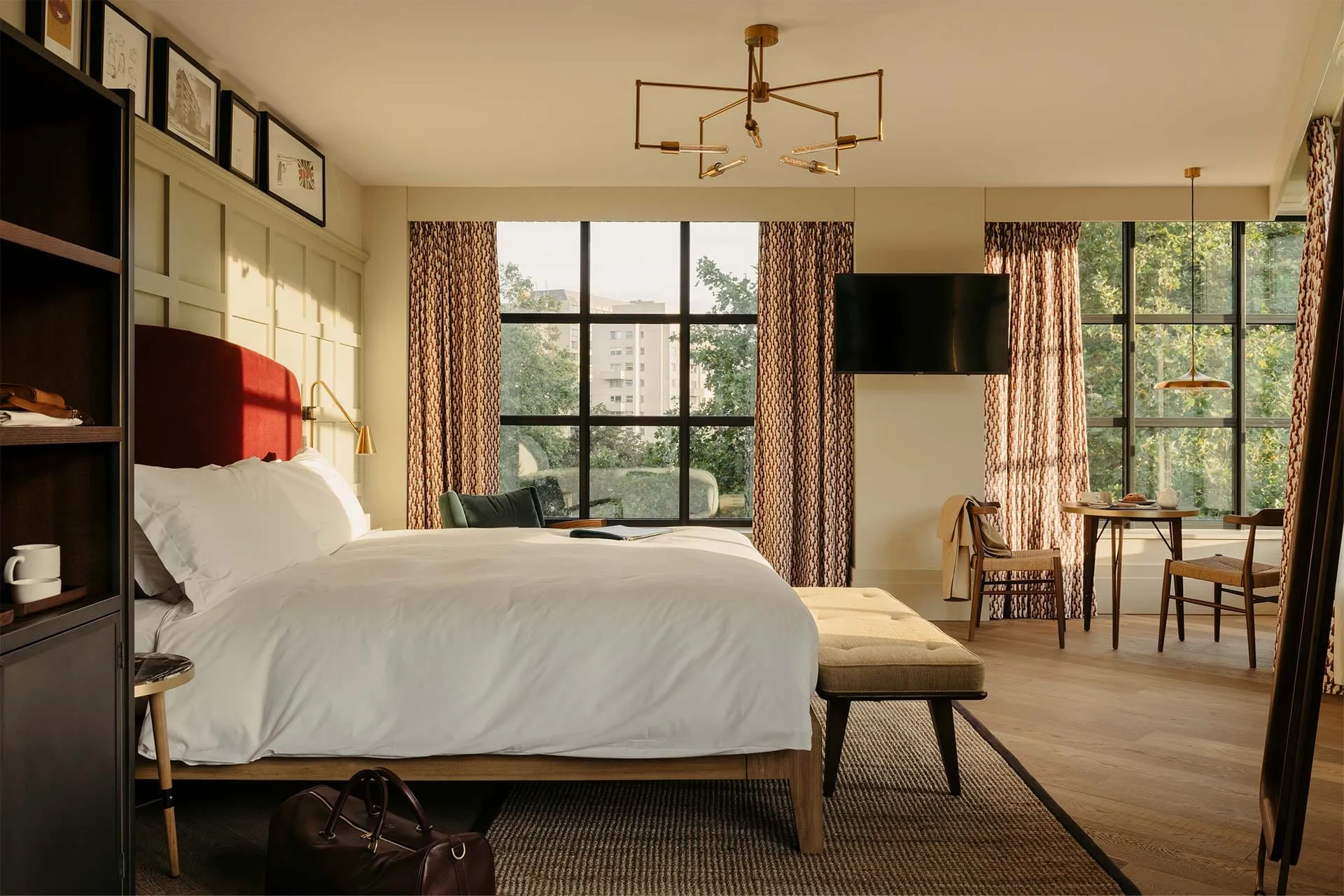 A room at The Hoxton, Southwark, with a burgundy velvet headboard, a gallery of framed prints above the bed and a seating and dining area by the window