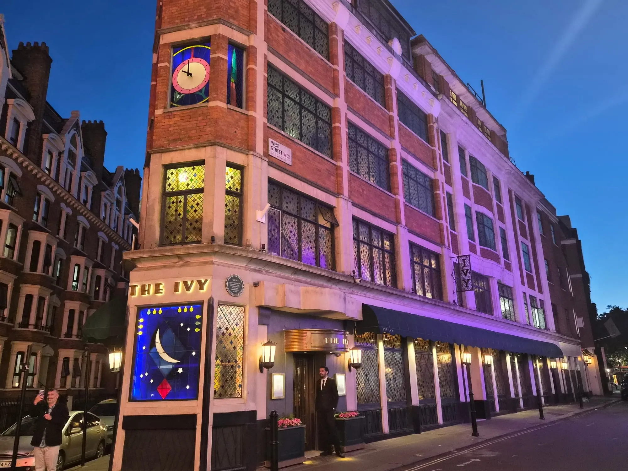 The Ivy's corner building on West Street at dusk, with the restaurant's name lettered on the stonework and a lit stained-glass window below.