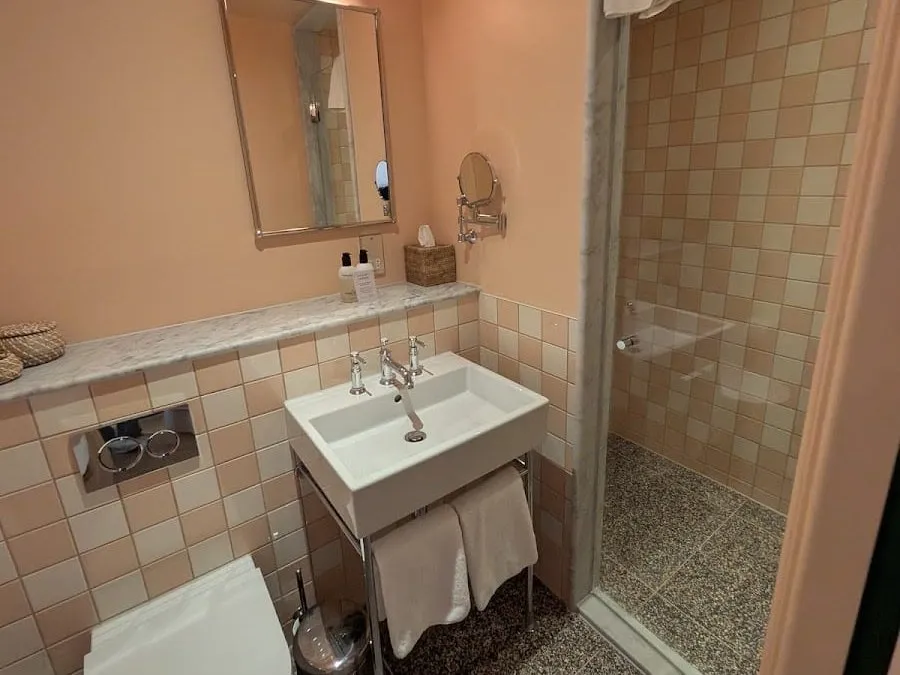 A bathroom at The July tiled in peach and cream squares, with a walk-in shower behind a glass screen and a white basin on a chrome stand