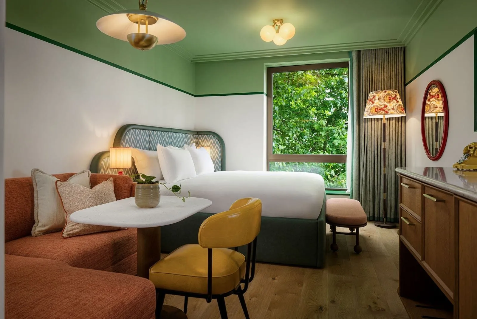 A room at The July with a green ceiling above white walls edged in green, a curved patterned headboard, an orange banquette and marble-topped table, a yellow leather chair and a window onto trees