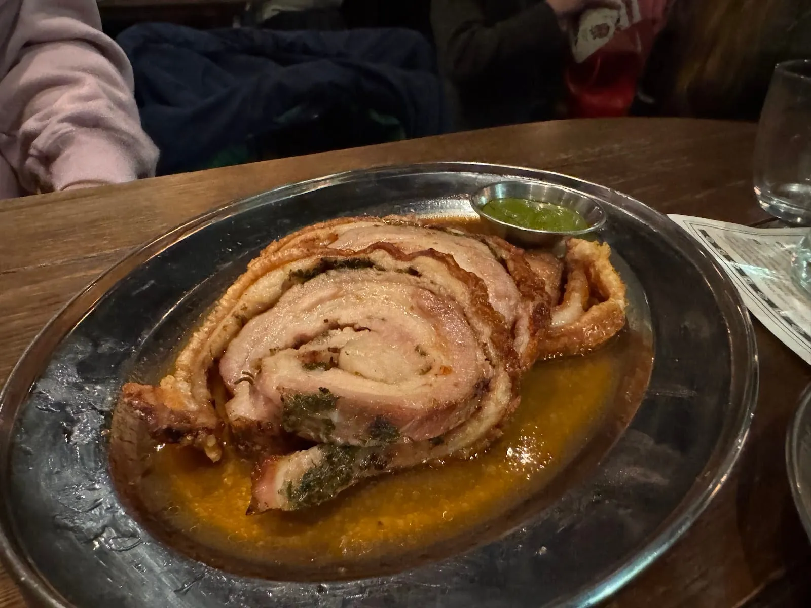 A rolled and sliced porchetta joint with crackling on a silver plate, with a small pot of salsa verde