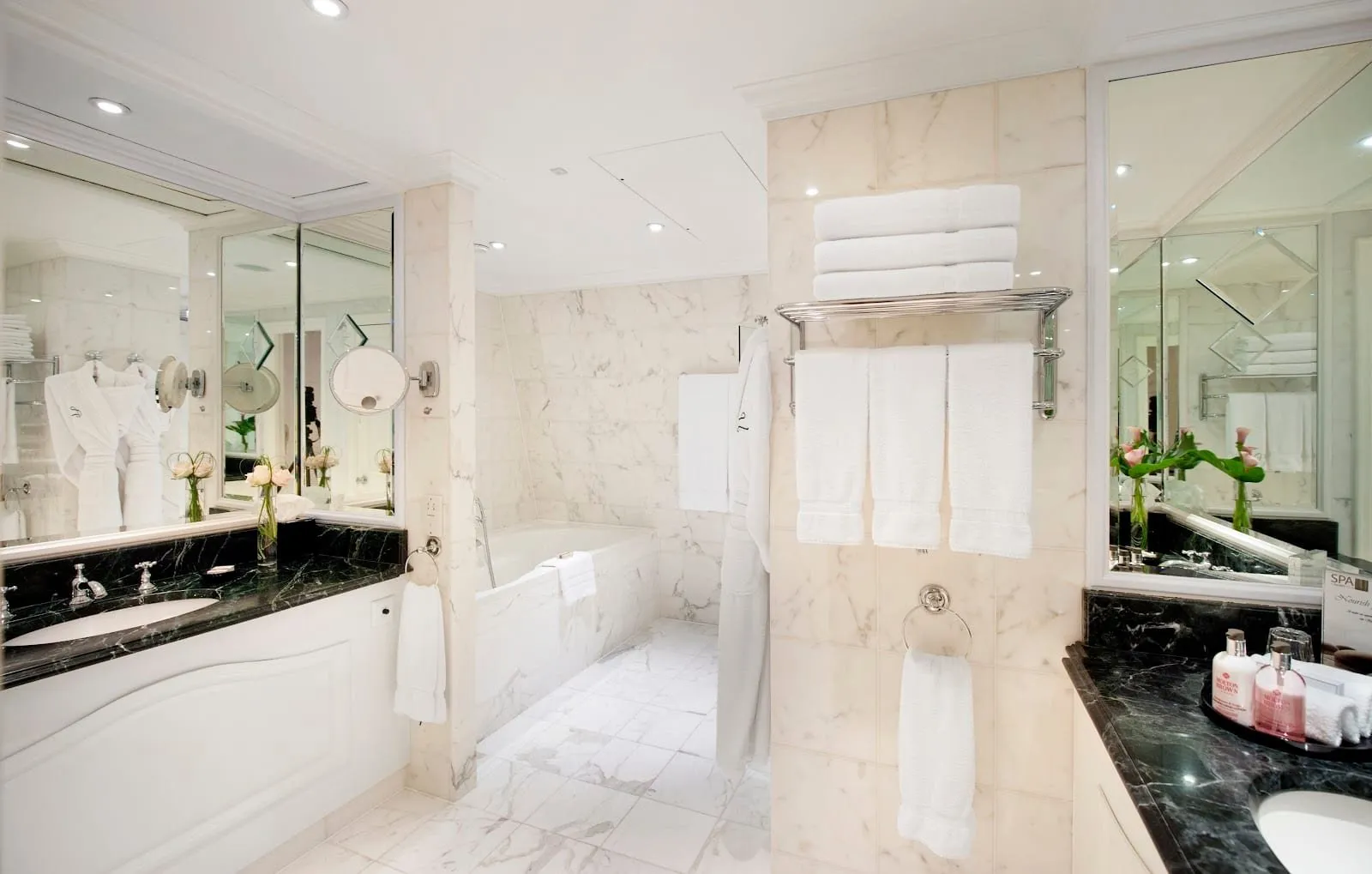 A white marble bathroom at The Landmark London with a bath set in an alcove, two basins on black marble counters facing each other, stacked towels and a bathrobe on a hook