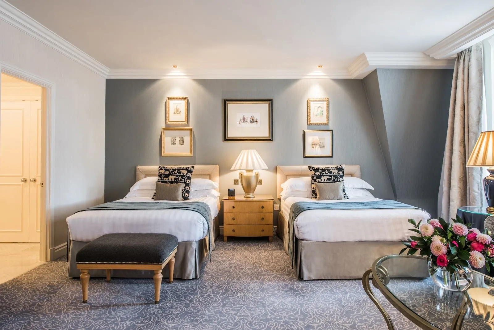 A room at The Landmark London with two double beds under cream padded headboards, a gold urn lamp on the chest between them, framed prints on a blue-grey wall and a glass table of pink dahlias