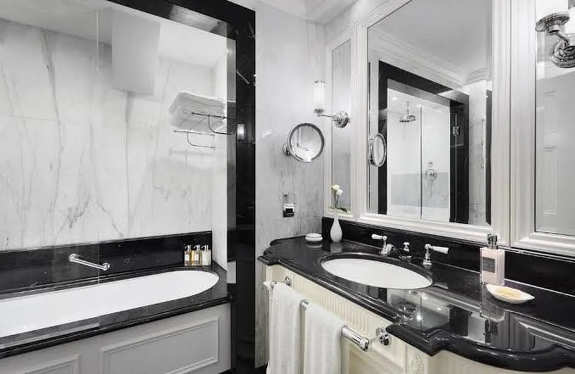 A Deluxe Room bathroom at The Lanesborough in white and black marble, with a bath behind a glass screen, a basin set in a black marble counter, and a separate walk-in shower reflected in the mirror