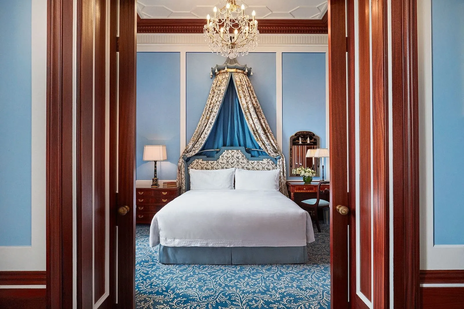 A Park Suite bedroom at The Lanesborough seen through mahogany double doors: a bed under a silk canopy crowned in blue and gold, pale blue panelled walls, a crystal chandelier and a blue patterned carpet