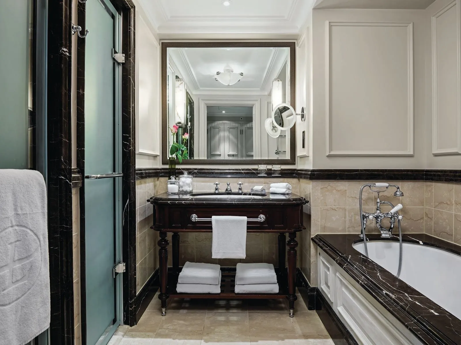 A bathroom at The Langham with a dark wood washstand under a large framed mirror, beige and dark brown marble, a bath with chrome telephone-style taps and a frosted glass door on the left