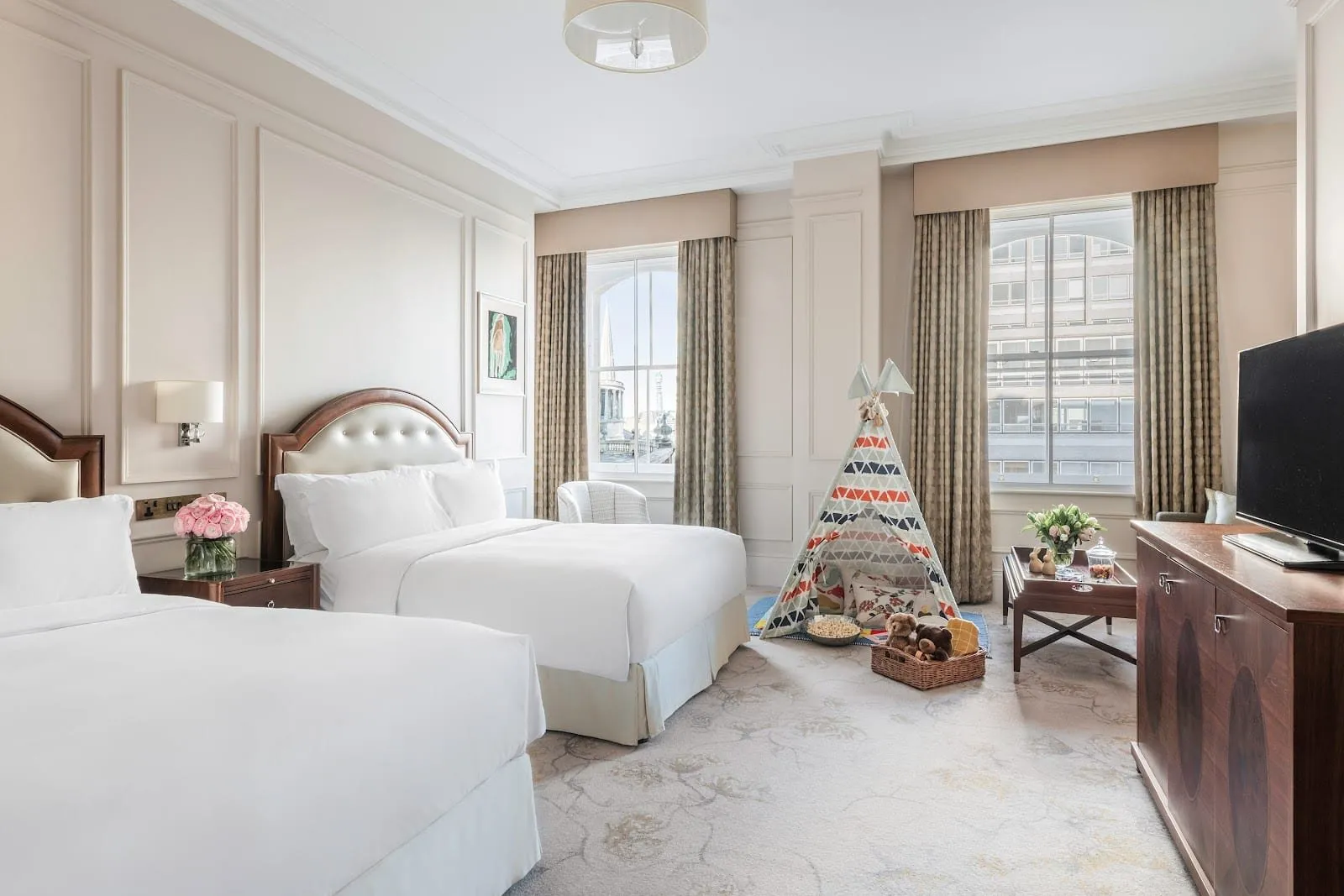 A family room at The Langham with two double beds under curved wooden headboards, a children's teepee with a teddy bear and popcorn by the window, and two tall sash windows, one looking out to the spire of All Souls Church