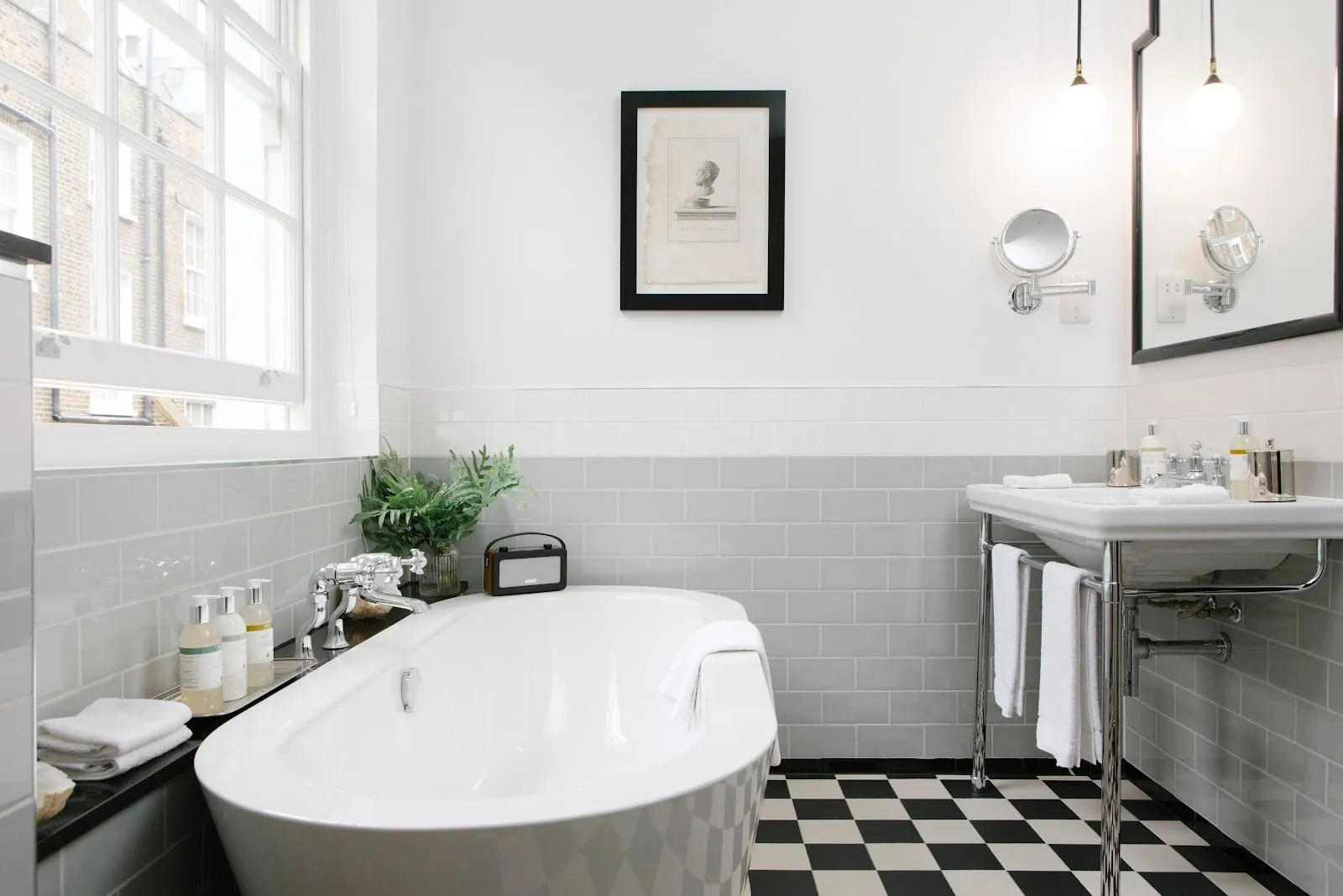 A bathroom at The Laslett with a freestanding white bath, grey metro tiles, a black and white chequerboard floor, a basin on a chrome washstand and a sash window onto brick houses