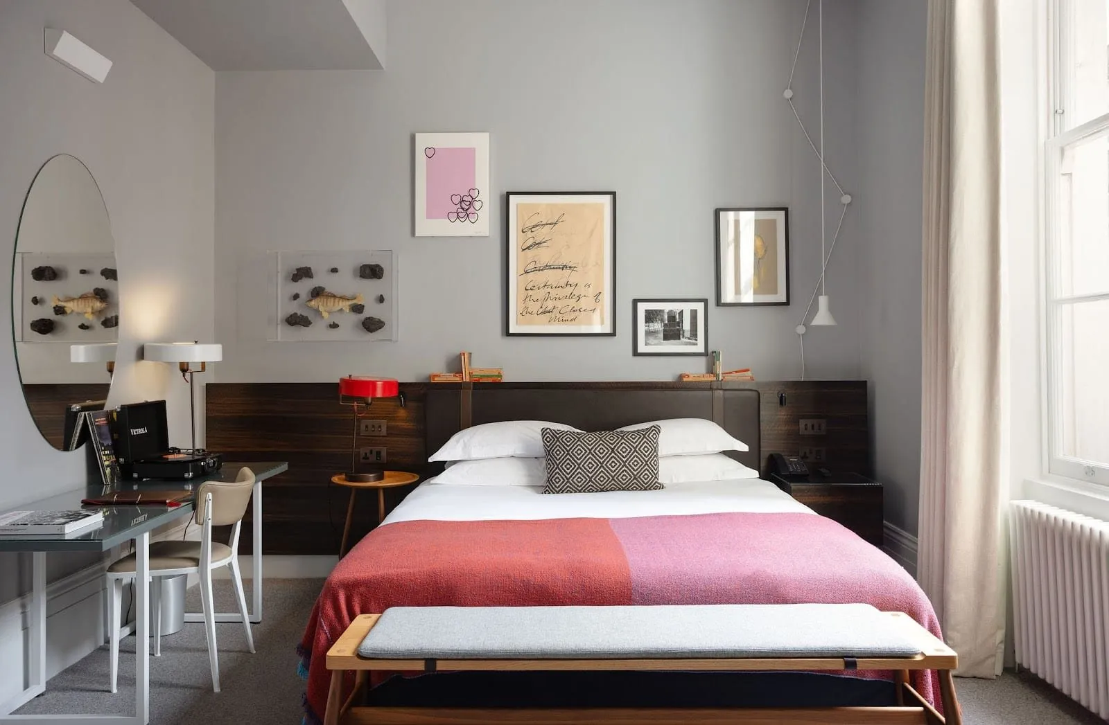 A double room at The Laslett with grey walls, a dark leather headboard set into a wood-veneer panel, a pink and red throw, a bench at the foot of the bed, a record player on the desk and a tall sash window