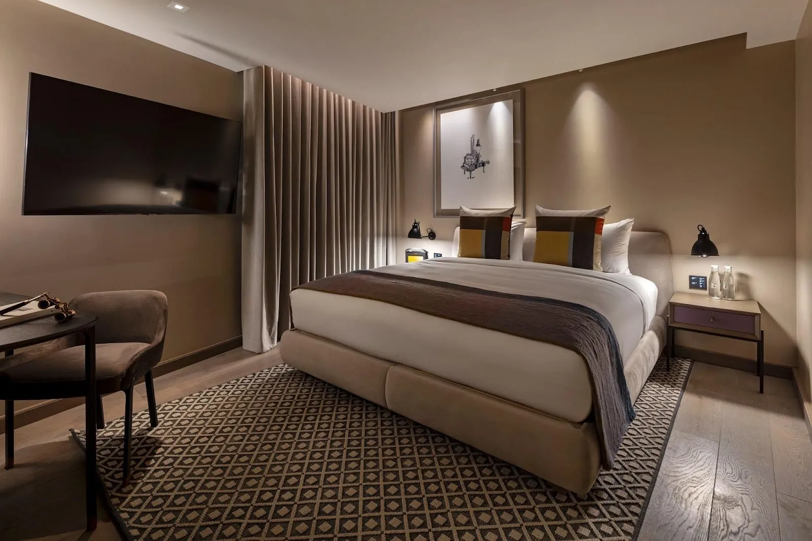 A guest room at the Londoner, with a wall-mounted television, a framed print above the bed and a desk beside the curtained window