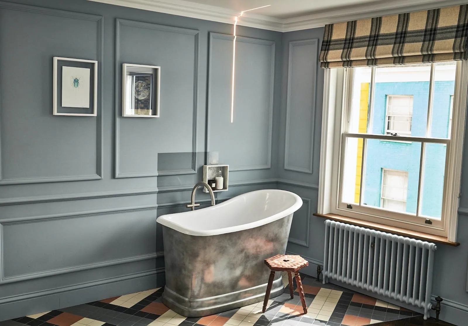 A bathroom at The Lost Poet with grey-blue panelled walls, a freestanding slipper bath with a pewter-finish outside, a patterned tiled floor, a wooden stool and a sash window facing a turquoise-painted house