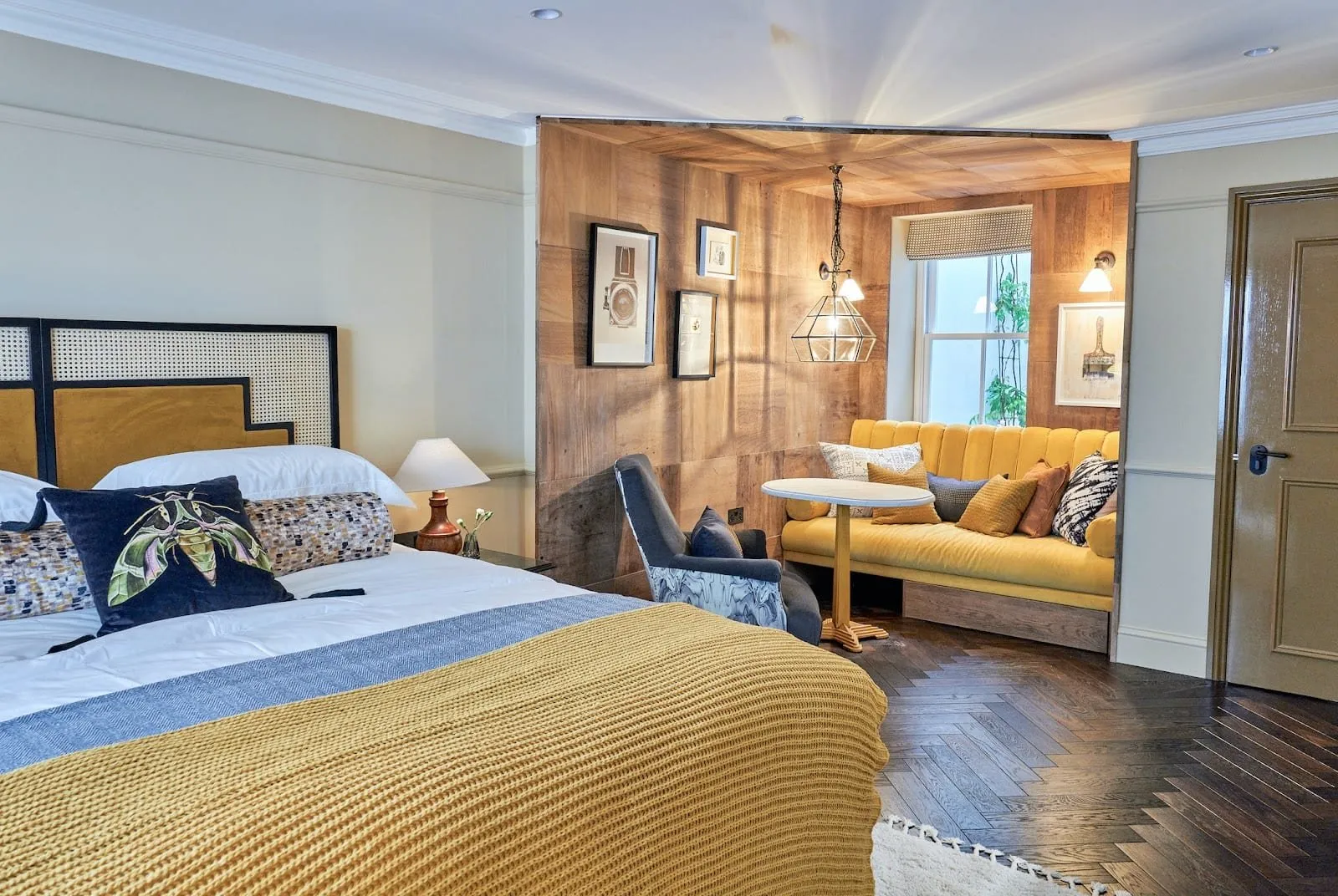 A room at The Lost Poet with a cane and mustard velvet headboard, a mustard knitted throw, herringbone floorboards and a wood-panelled nook with a yellow velvet banquette and a small round table by a window