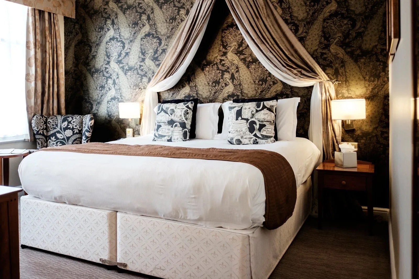 A double room at The Mad Hatter with peacock-pattern wallpaper, a draped fabric canopy over the bed, a floral wingback chair and a patterned divan base
