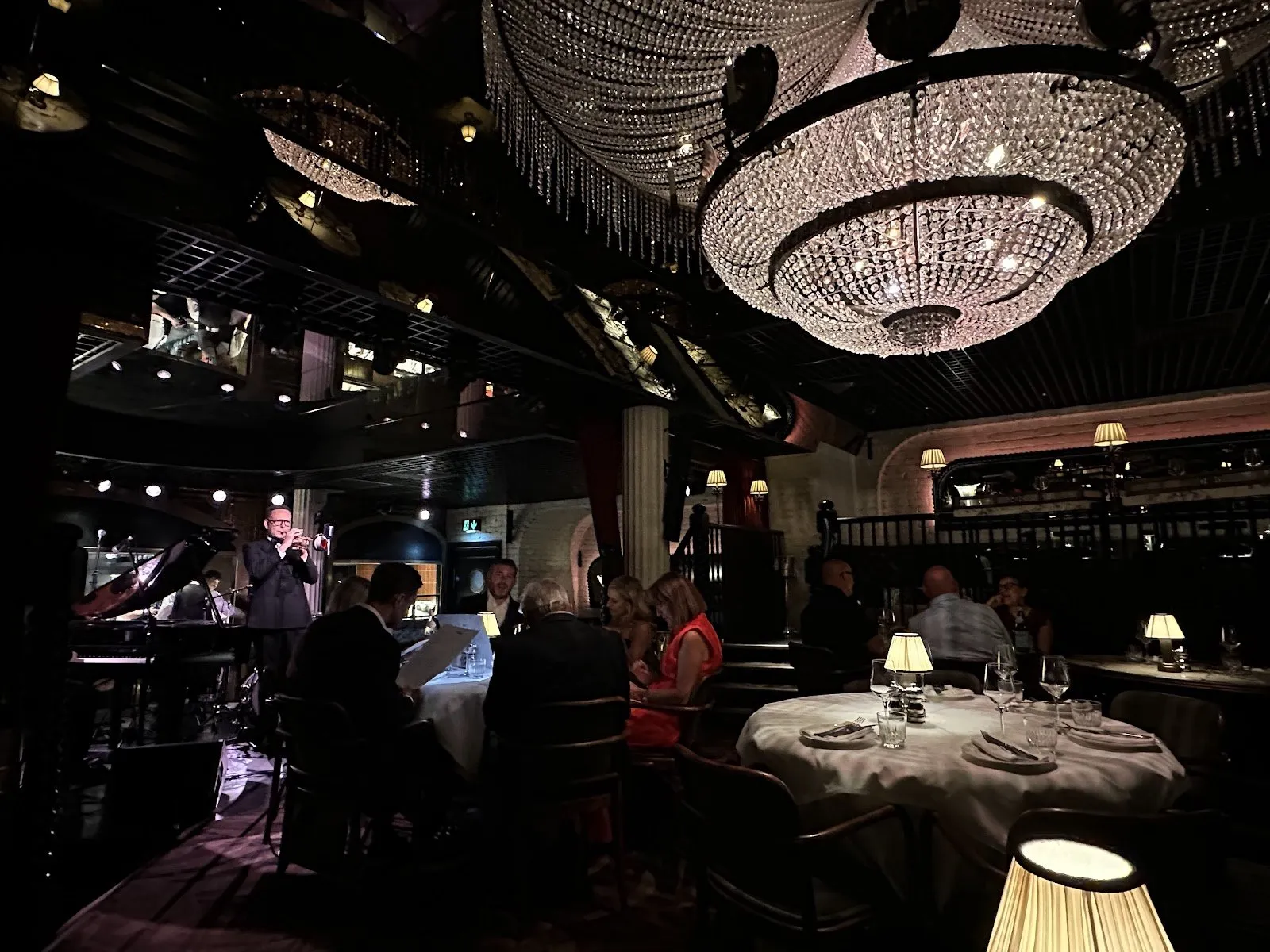 A trumpeter performs on the brasserie stage at The Maine under a large crystal chandelier, with diners at nearby tables