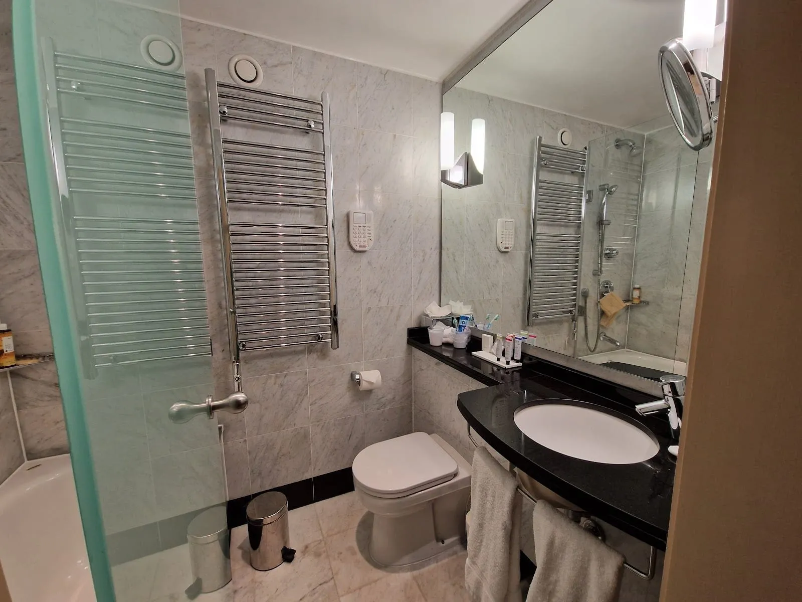 A grey marble bathroom at The Mandeville with two chrome heated towel rails, a black granite vanity with an oval basin, and a bath with a hand shower reflected in the wide mirror