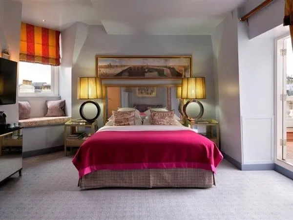 A top-floor room at The Mandeville with a magenta blanket on the bed, gold table lamps, a cushioned window seat under a red and yellow striped blind, and a glazed door on the right opening on to a terrace
