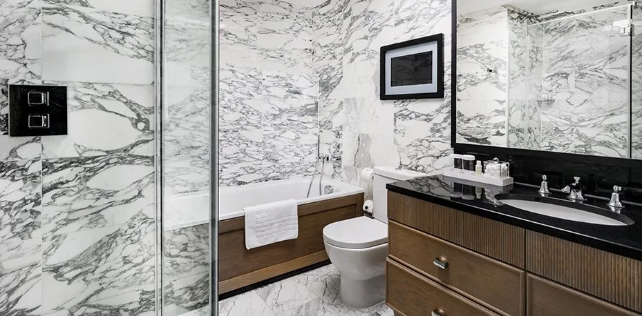 A bathroom at The Marylebone lined in grey-veined white marble, with a glass shower on the left, a wood-panelled bath beyond and a black-topped vanity with a ribbed wooden cabinet