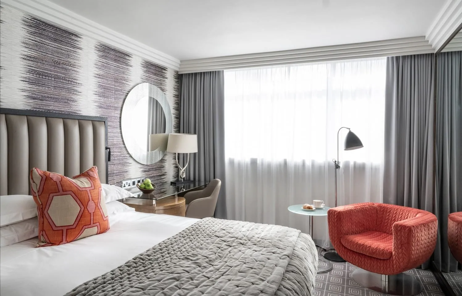 A bedroom at The Marylebone with a grey leather channelled headboard against purple-streaked wallpaper, a coral swivel armchair, a round mirror over the desk and a wide window behind sheer curtains