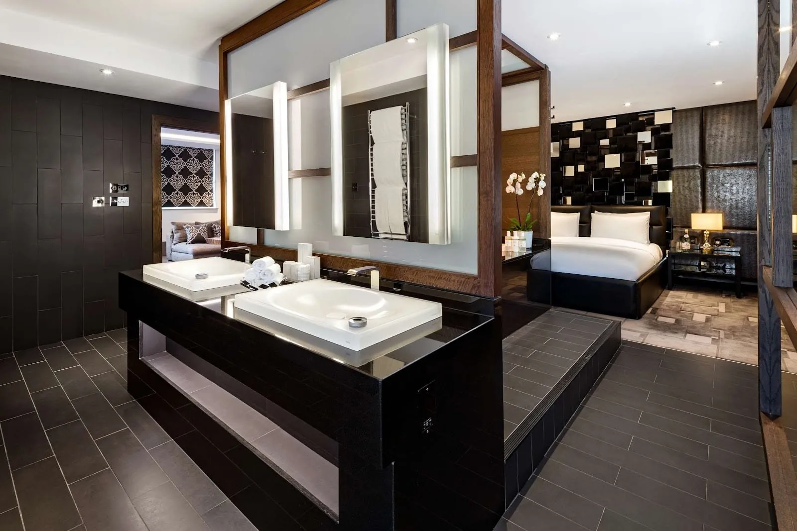 A May Fair suite bathroom in dark slate tiles with twin white basins on a black counter, open to the bedroom on one side and a walk-in shower on the other