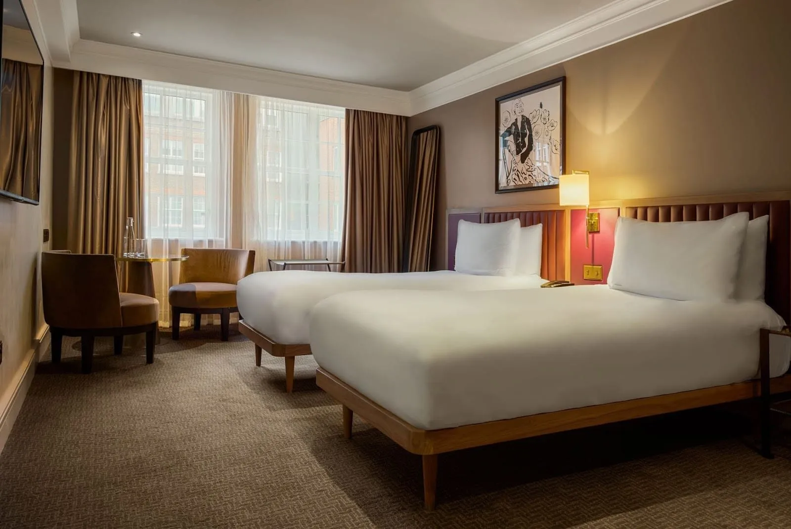 A twin room at The May Fair with two single beds on wooden frames under padded leather headboards, two tan armchairs at a round table and a window onto red-brick buildings