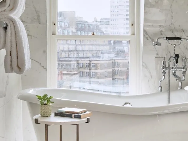 A Mayfair Townhouse bathroom with a white slipper bath under a sash window, marble walls, a chrome telephone-style mixer and a small side table with books