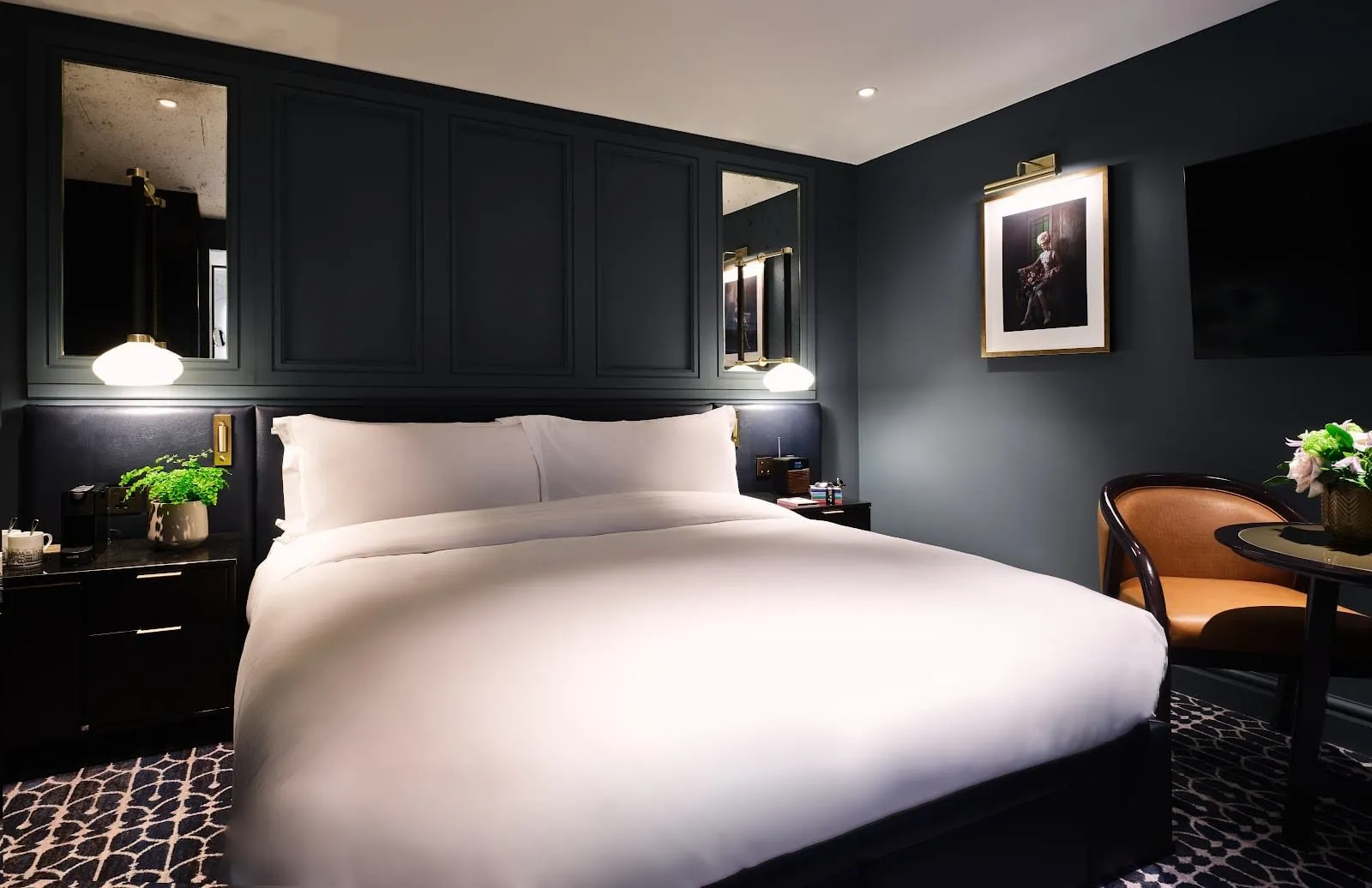A Mayfair Townhouse bedroom with charcoal panelled walls, a navy leather headboard with brass reading lights, a tan leather chair at a small round table and a framed portrait photograph