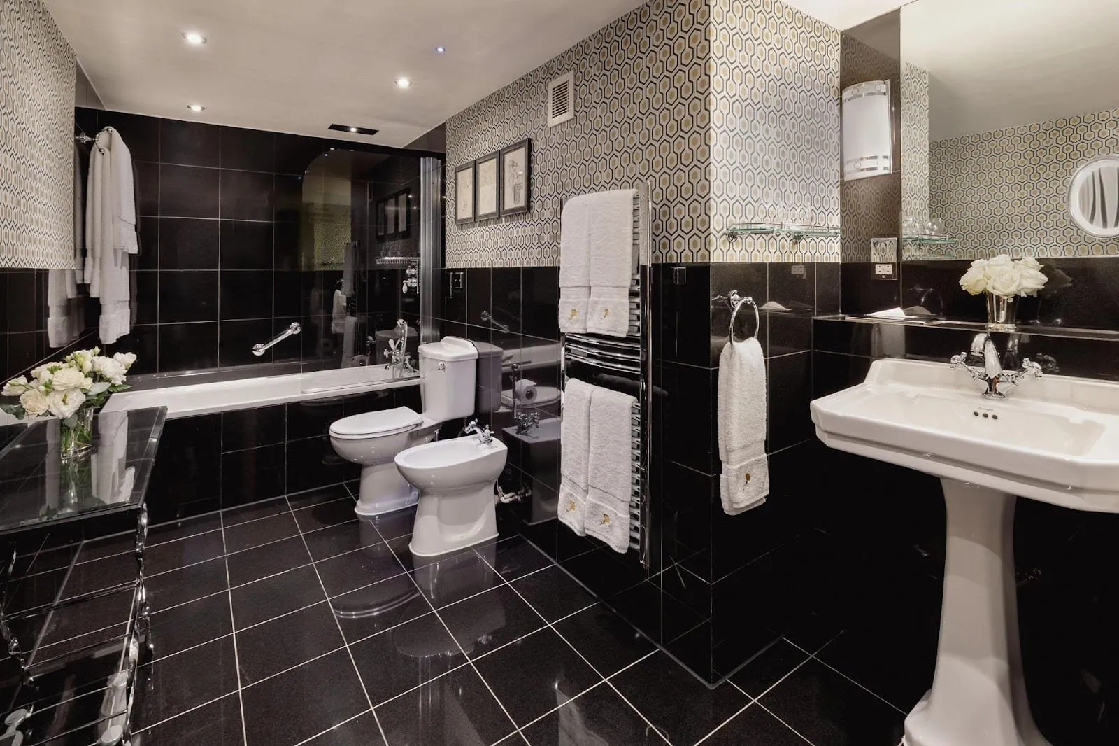 A Milestone bathroom in black granite tiles and geometric wallpaper, with a bath behind a glass screen, a bidet, a pedestal basin and embroidered towels on a heated rail