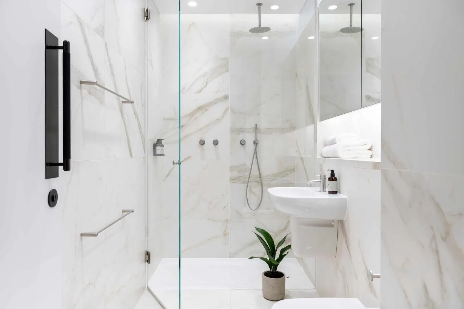 A bathroom at The Moorgate by Cove in white marble-effect tiles, with a walk-in rain shower behind a glass door, a wall-hung basin and a towel shelf