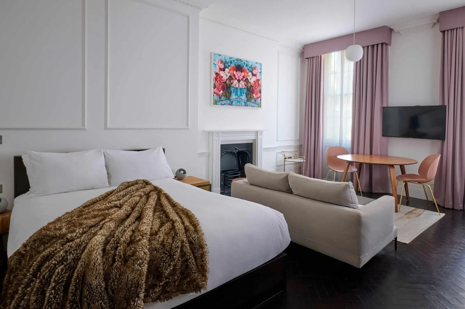 An apartment at The Moorgate by Cove with white panelled walls, a period fireplace, a bed with a fur throw, a grey sofa, a round table with pink chairs, dark herringbone floors and dusky pink curtains