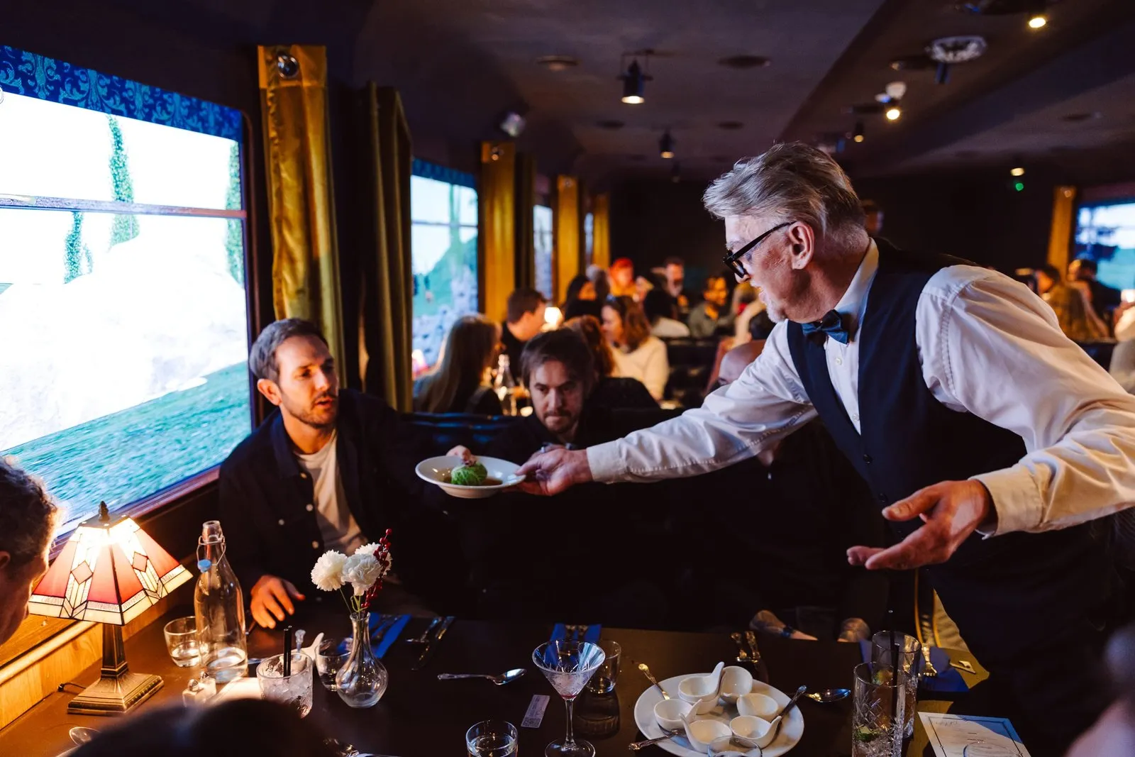 A waiter in a waistcoat and bow tie serves a dish to diners in the train-carriage dining car, with a landscape projected in the windows