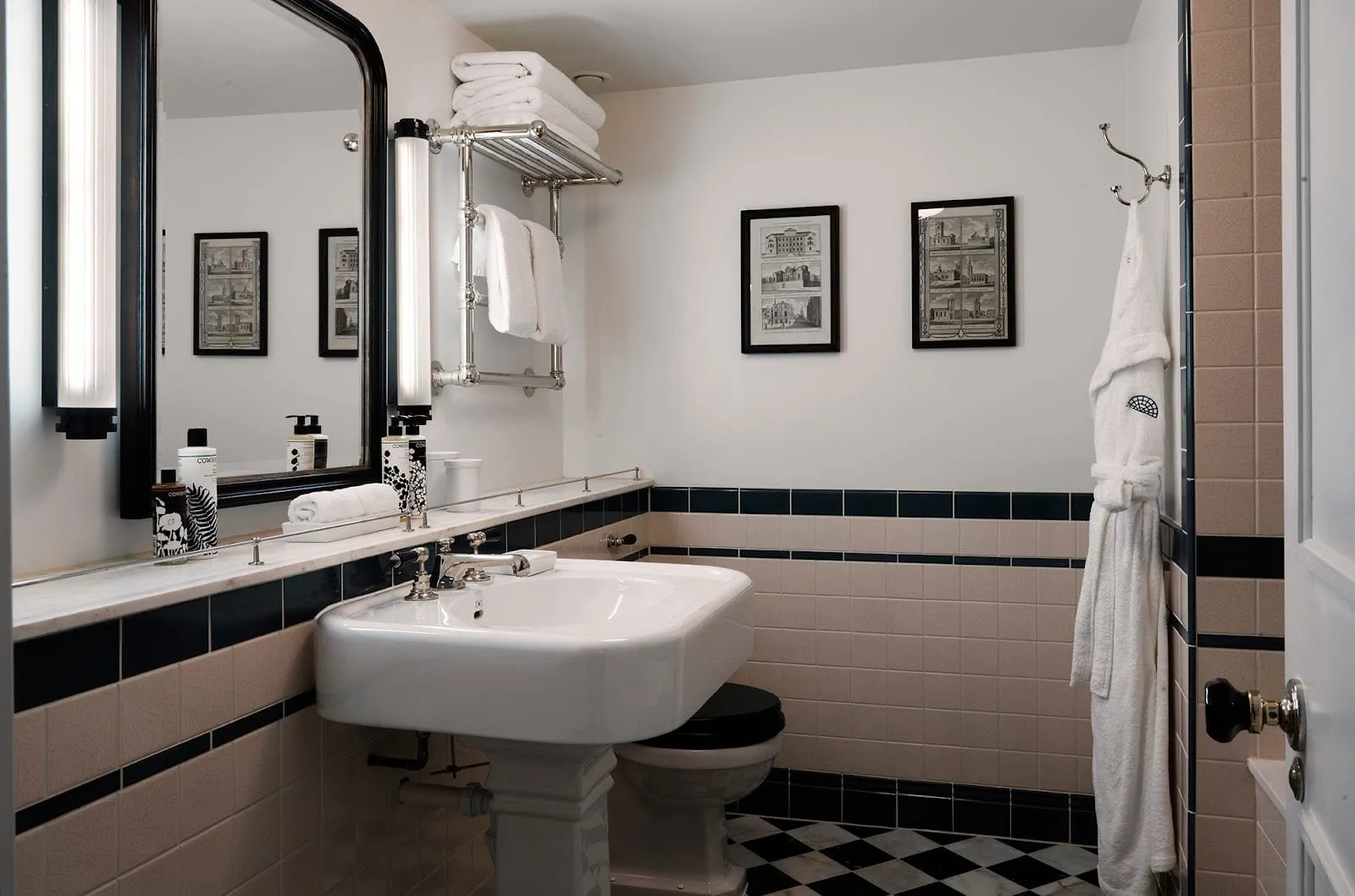 A bathroom at The Ned with a white pedestal basin, cream tiles banded in black, a chequered marble floor, a nickel towel rack and framed engravings of City buildings