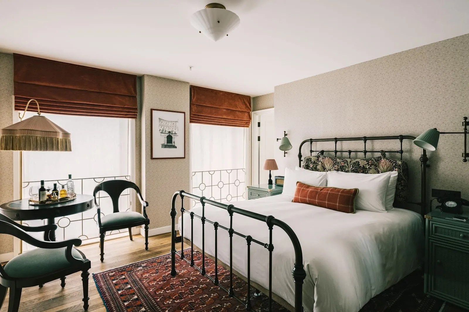 A double room at The Ned with a dark iron bedstead, an Arts and Crafts patterned headboard, a red Turkmen rug on oak boards, rust velvet blinds at two tall windows and a small round table with a drinks tray
