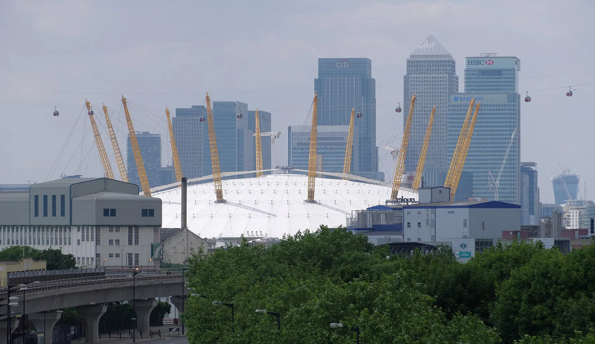The O2's white dome and yellow masts with cable car gondolas crossing in front of the Canary Wharf towers, seen from the Royal Docks side with a DLR viaduct in the foreground