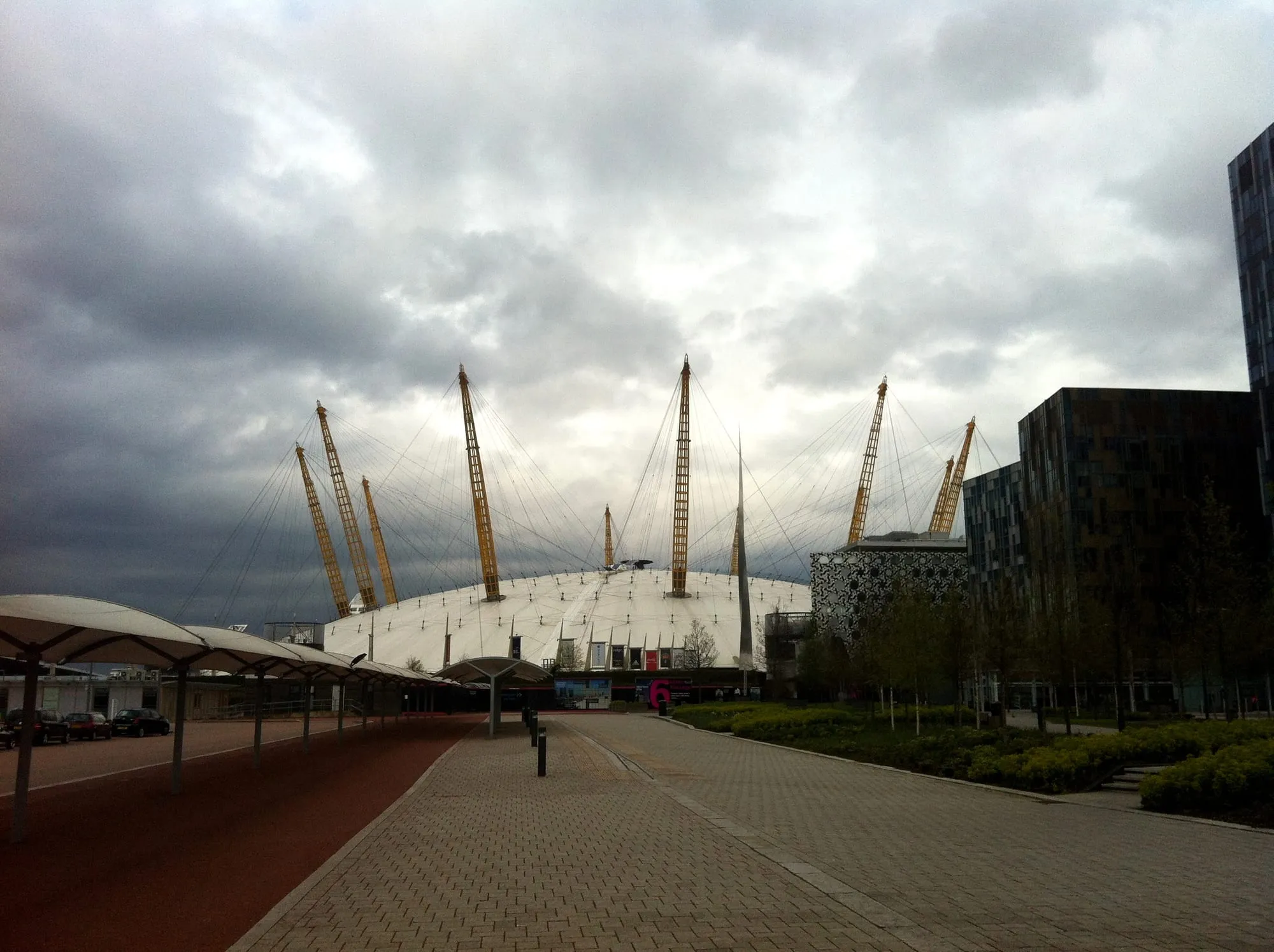 The O2's white dome and yellow support masts seen from the empty forecourt under a grey sky