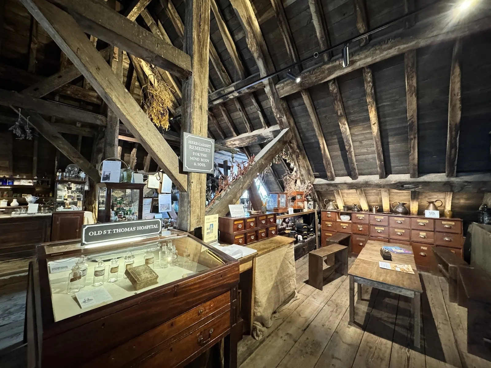 The herb garret under the church's bare timber roof beams, with apothecary drawers, glass cases of medicine bottles, dried herbs hanging from the rafters and a sign reading Old St Thomas' Hospital