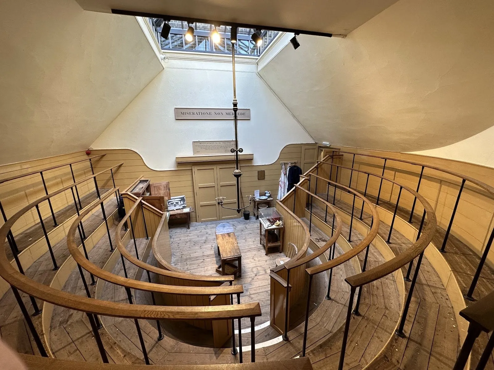The Old Operating Theatre from the top tier: curved wooden standing rails in rising rows round a plain wooden operating table, under a skylight, with a sign reading Miseratione non mercede over the double doors