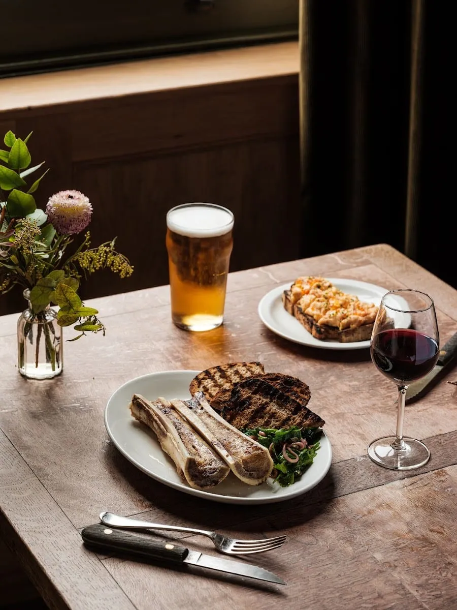 Grilled bone marrow with toast and herbs, alongside a small plate of food and a glass of red wine, on a wooden table at The Pelican