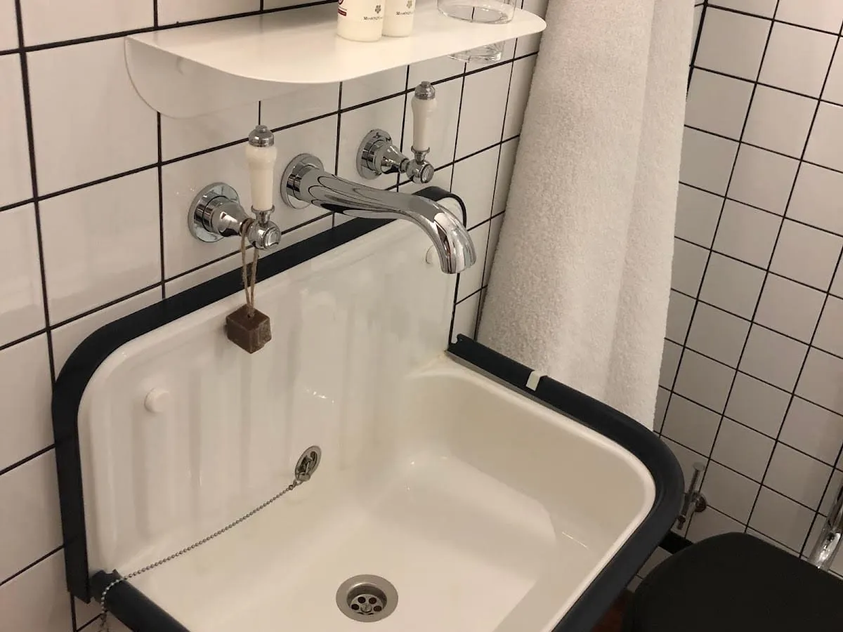 An enamel utility-style basin with brass-and-porcelain taps and a soap on a rope, against white tiles with black grout, at The Pilgrm