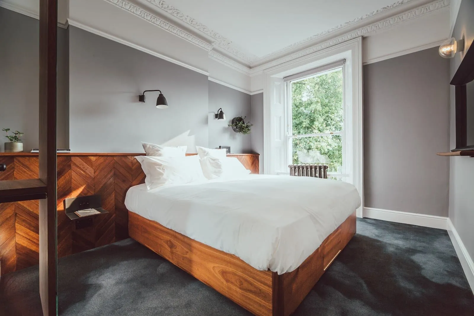 A double room at The Pilgrm with a full-height sash window onto trees, ornate Victorian cornicing, herringbone wood panelling behind a walnut bed and grey walls
