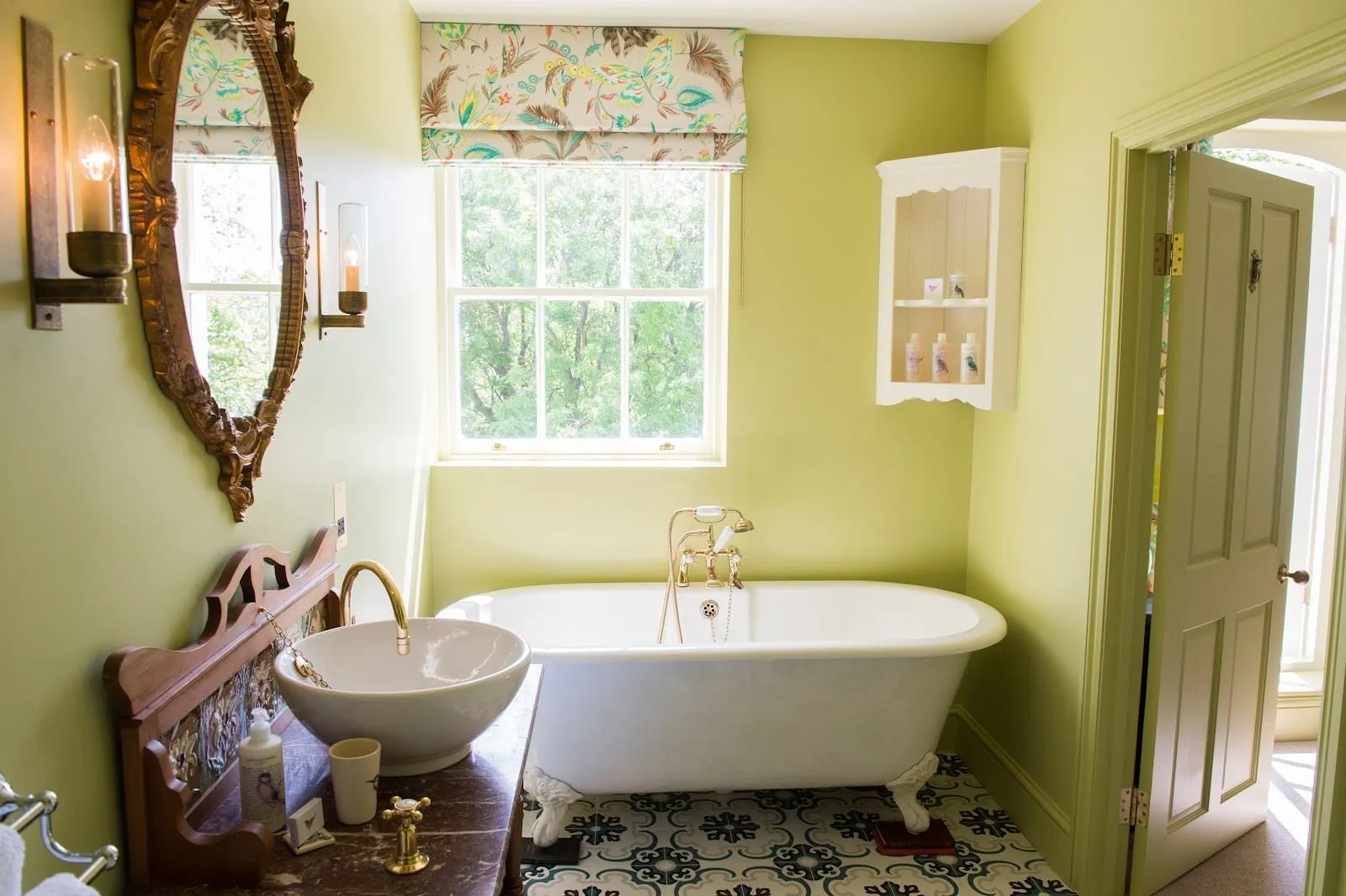 A bathroom at The Portobello Hotel with lime-green walls, a white roll-top bath on claw feet with brass taps, a bowl basin on an antique wooden washstand, an ornate gilt mirror and a sash window onto trees