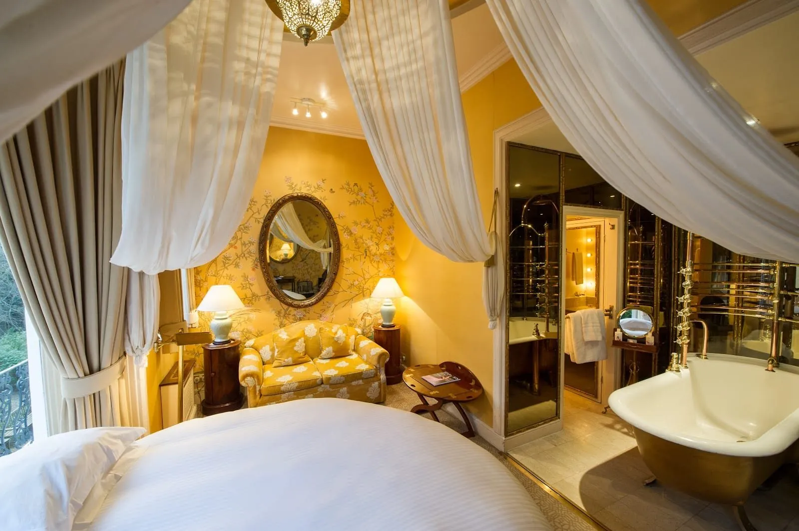 A room at The Portobello Hotel with a round bed under white muslin drapes, yellow hand-painted wallpaper of birds and blossom, a yellow sofa, an oval gilt mirror and a roll-top bath standing in the bedroom beside the doorway to the washroom