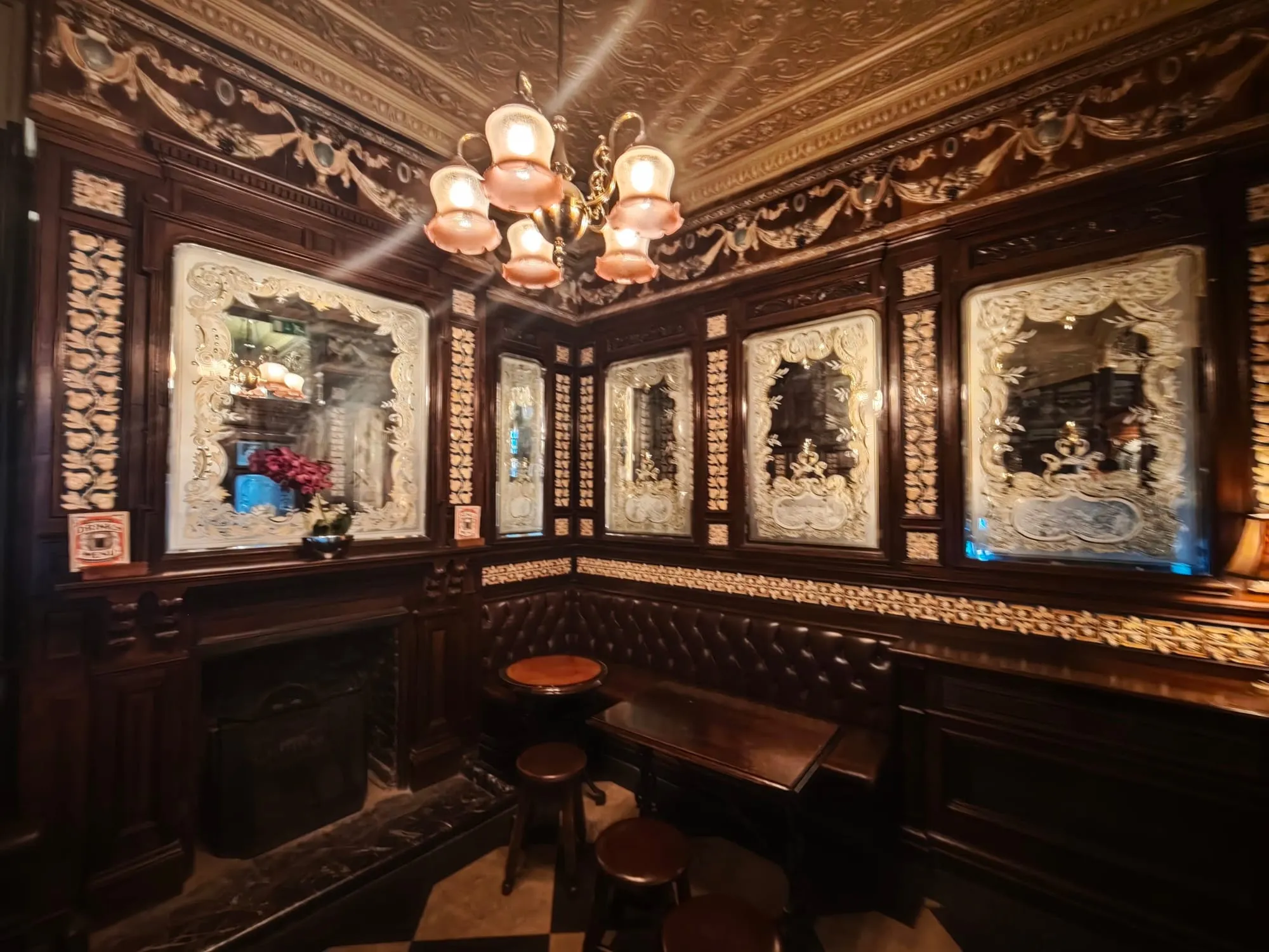 A wood-panelled corner of The Princess Louise with a leather banquette, small tables and etched mirrors under a patterned ceiling