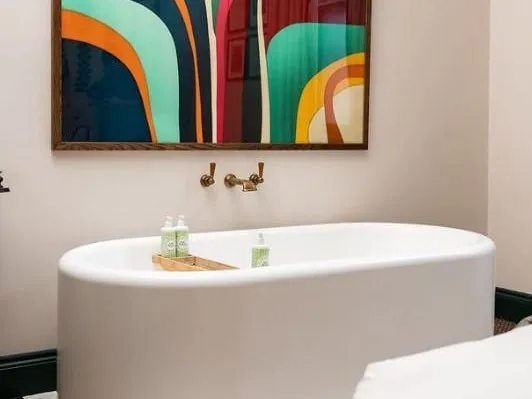 A white freestanding bath with brass wall taps under a colourful abstract print, standing in the bedroom at The Princess Royal with the corner of the bed in view